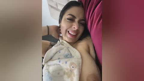 Media: A video of a smiling, dark-haired woman with medium skin tone, topless, lying on a pink pillow, covered in a white towel with cartoon characters, in a dimly lit room.