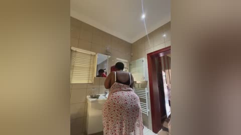 Video of a dark-skinned woman in a floral dress, standing in a beige-tiled bathroom with a mirror, towel rack, and window blinds.