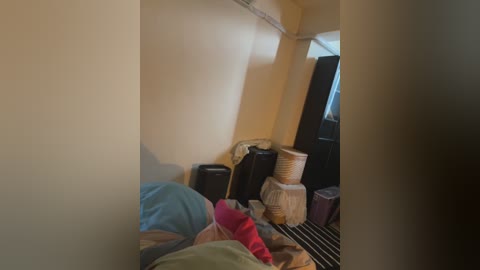 A video of a cluttered, dimly-lit room with beige walls, featuring a person in a green shirt, a pile of clothes, and several large black suitcases and a woven basket.