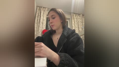 A video of a young woman in a black robe, focused on a smartphone, in a dimly lit room with beige curtains.