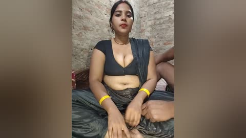 A video of a South Asian woman with medium skin tone, wearing a black sari and a black bra, sitting on a bed with exposed brick walls. She has a confident expression, yellow bangles, and a gold necklace.