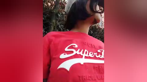 Video of a young person with medium-length dark hair wearing a red \"SuperT\" sweatshirt, partially obscured by a blurred background featuring a chain-link fence and green foliage.