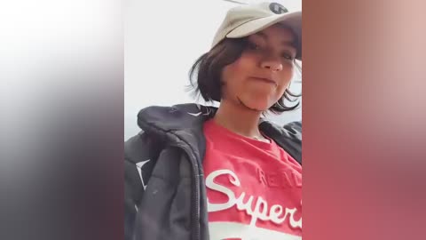 Video of a young Asian woman with short dark hair, wearing a beige baseball cap and a red \"Super\" t-shirt, standing in a blurred, indoor setting.