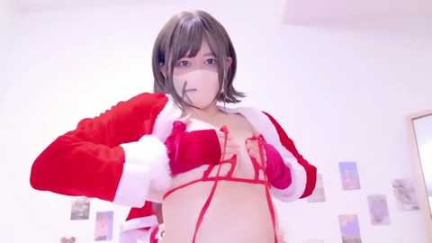 Video of a young woman with short, dark hair, wearing a red and white Santa-themed bra, with a surgical mask, standing in a bright, minimalist room with white walls and a mirror.