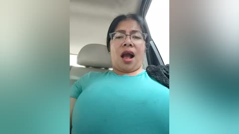 Video of a middle-aged Asian woman with glasses, wearing a tight turquoise shirt, mouth open in surprise or excitement, seated in a car.