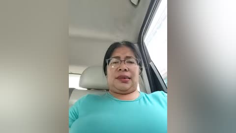 Video of a middle-aged woman with short black hair, wearing glasses and a turquoise shirt, sitting in a car with a beige headrest and window beside her.