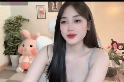 A video of a young Asian woman with long black hair, fair skin, and red lipstick, wearing a blue spaghetti-strap top, standing in a cozy room with plush toys, plants, and framed photos.
