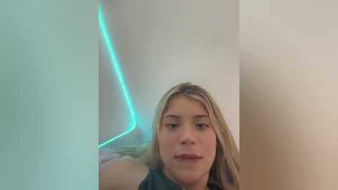 Video of a young woman with straight blonde hair, fair skin, and wearing a dark top, smiling slightly. Background features a glowing cyan LED strip and a white wall.