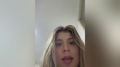 A video of a young woman with light skin, blonde hair, and slightly parted lips, taken from a low angle, showing her face and upper chest. The background is blurred, with a white wall and a hint of a ceiling.