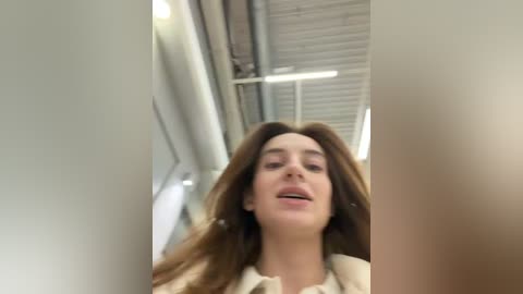 Media: A video of a young woman with long, straight brown hair, wearing a beige blouse, captured from a low angle in a modern, brightly lit room with white walls and a window with horizontal blinds.
