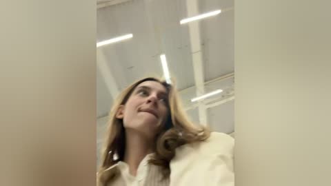 Media: Video of a young woman with long, wavy blonde hair, wearing a beige coat, standing in a brightly lit, modern indoor space with white walls and ceiling.