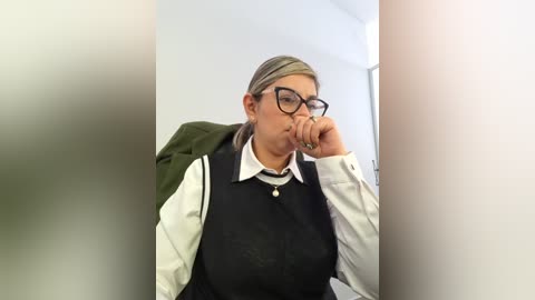 Video of a woman with glasses, black vest, and white blouse, sitting with hand to mouth, in a blurred, possibly office setting.