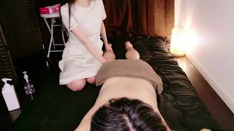 Video of a woman in a white dress kneading a man's back on a dark green mat in a dimly lit room with a white lamp.