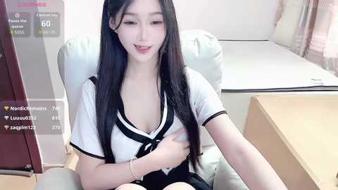 Video of an Asian woman with long black hair, wearing a white sailor uniform, seated in a chair, smiling, in a modern room with wooden furniture.