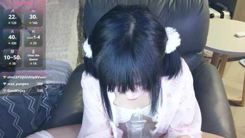 Video of an Asian woman with straight black hair, wearing a white nightgown, kneeling on a black leather chair, drinking from a glass, in a dimly lit room.