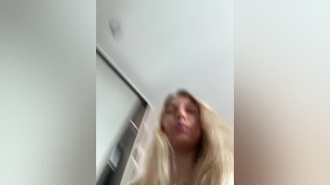 A blurry video shows a blonde woman with long hair, making a pouty face, standing in a white-walled room with a metal-framed mirror.