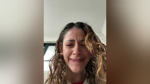 Video of a woman with curly, shoulder-length hair, smiling broadly, wearing a light-colored top, indoors with a blurred window in the background.