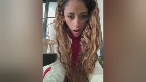 Video of a young woman with light brown skin and curly, wavy, ombre hair, wearing a red shirt, leaning over a white couch, looking surprised, indoors with a window and plants in the background.