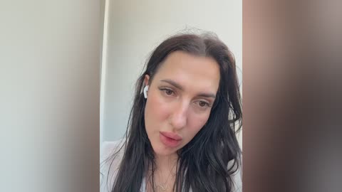 Video of a young woman with long, straight black hair and fair skin, wearing white headphones, against a blurred background.