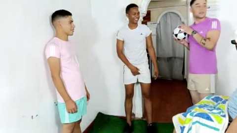 Video of four young men in casual sportswear, three in white and one in purple, standing in a brightly lit room with a soccer ball.