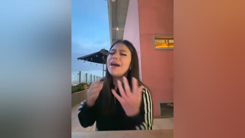 Video of a young woman with long black hair, wearing a black jacket with white stripes, making a surprised or shocked face with her hands raised. Background shows a pink building and a clear blue sky.