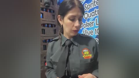 Video of a woman in a dark, gray uniform with a yellow badge, standing in a dimly lit room with a blue wall displaying white text and a wooden cabinet with shelves.
