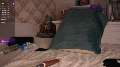 Video of a messy bedroom with a teal quilt, scattered clothes, a laptop, and a man partially visible in the background.