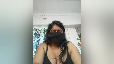 Video of a South Asian woman with medium brown skin, wearing a black face mask, black tank top, and long black hair. Background includes white walls and floral tapestry.