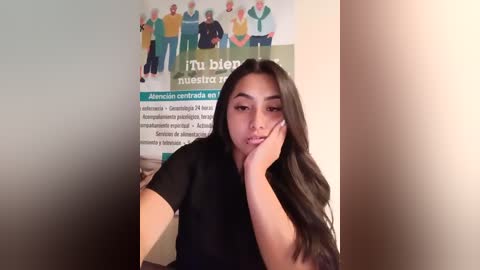 Video of a young woman with long black hair, wearing a black t-shirt, sitting in front of a \"Mental Health\" poster.