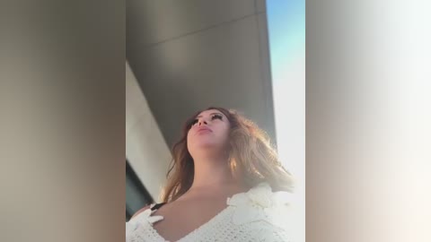 Video of a fair-skinned woman with long, wavy brown hair, wearing a white, crocheted top, standing in a sunlit, modern building corridor. The image is slightly blurred, with sunlight creating a soft glow.