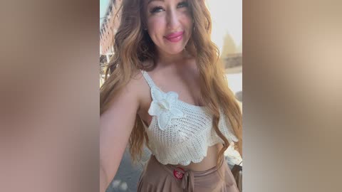 A video of a smiling woman with long, wavy brown hair, wearing a white lace crop top with a floral design and high-waisted beige skirt. The background shows a blurred outdoor setting with a brick building.