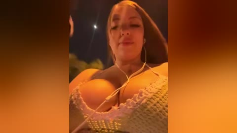 Video of a fair-skinned woman with long brown hair, wearing a knitted sweater, smiling while listening to music with earphones, under a clear night sky with a full moon.