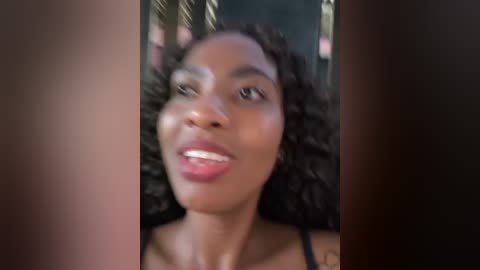 A video of a Black woman with curly hair, wearing a black top, smiling, with blurred background suggesting an outdoor setting.