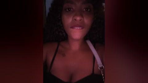 Video of a young Black woman with medium skin tone, curly hair, and a light pink shoulder bag, wearing a black spaghetti-strap top, in dim lighting.