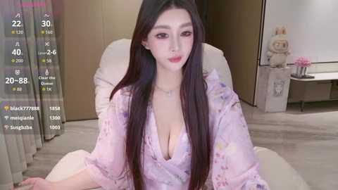 Video of a young Asian woman with long black hair, fair skin, and large breasts, wearing a pink floral robe, sitting on a beige chair in a modern, minimalistic bedroom.