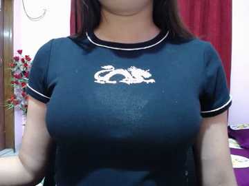 Video of a woman with medium breasts wearing a dark blue, short-sleeved t-shirt with white dragon logo. She has light skin and shoulder-length brown hair. Background includes red curtain and floral arrangement.