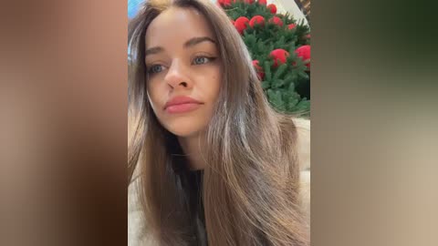 A video of a young Caucasian woman with long, straight, light brown hair and blue eyes, wearing a black top. Background features a vibrant arrangement of red tulips and green foliage.