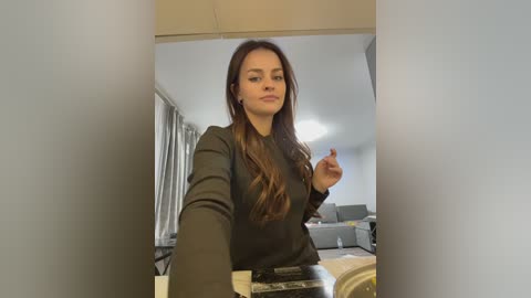 Video of a young woman with long, wavy brown hair, wearing a dark green sweater, holding a pen in a modern, minimalist living room with light-colored walls and a sofa in the background.