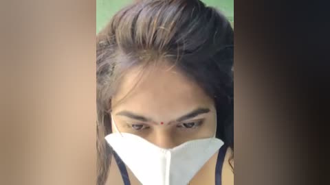 Video of a young woman with medium-brown skin, wearing a white surgical mask, her long, dark brown hair partially tied up, and a red bindi on her forehead, focusing intently on a task.