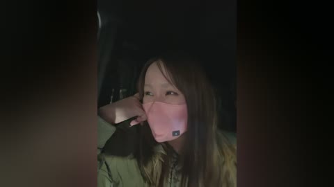 Video of a woman with long brown hair, wearing a pink face mask, sitting in a dimly lit car, eyes half-closed, looking tired.