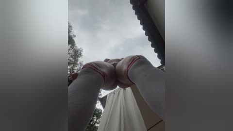 Video of a woman's legs wearing white knee-high socks and red-striped socks, standing under an umbrella in a cloudy outdoor setting with trees and a building in the background.