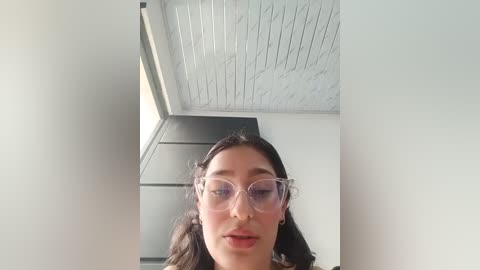 A video of a young woman with fair skin, dark hair, and clear glasses, standing in a modern room with white walls, a white ceiling, and a black-framed window.