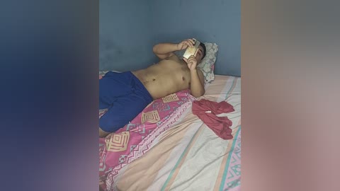 Video of a shirtless man in blue shorts, lying on a bed with a pink-patterned blanket, holding a phone to his ear, in a simple bedroom with blue walls.