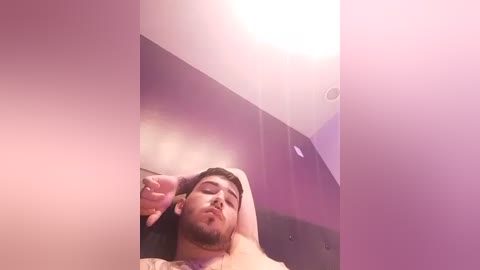 A video of a man with a trimmed beard, lying shirtless on a bed, arms above head, in a dimly lit room with purple walls and a ceiling light.