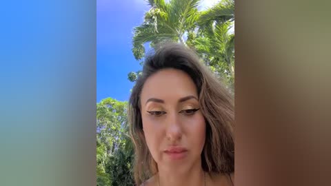Video of a young woman with long brown hair and light skin, wearing a light-colored top, standing against a lush green palm tree against a clear blue sky.