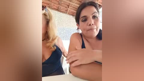 Video of a woman with light brown skin, dark hair, and medium breasts in a black top, sitting at a table, with a blurred, partially nude blonde woman in the background.