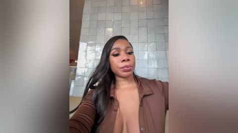 Video of a Black woman with long, straight black hair, wearing a brown jacket, posing in a modern, tiled bathroom.