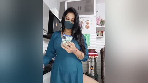 Video of a woman with long black hair, wearing a teal long-sleeved dress, blue surgical mask, and green-patterned shirt, holding a smartphone, in a modern kitchen with white cabinets, red stools, and floral decorations.