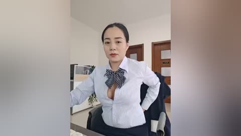 Video of an Asian woman in a professional setting, wearing a light blue blouse with a bold black and white striped bow tie, sitting at a desk, with a blurred background of office furniture.