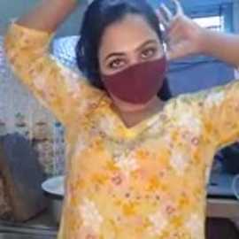Video of a woman with medium skin tone, wearing a yellow floral dress, maroon face mask, and a maroon headband, posing indoors with hands up, blurred background.
